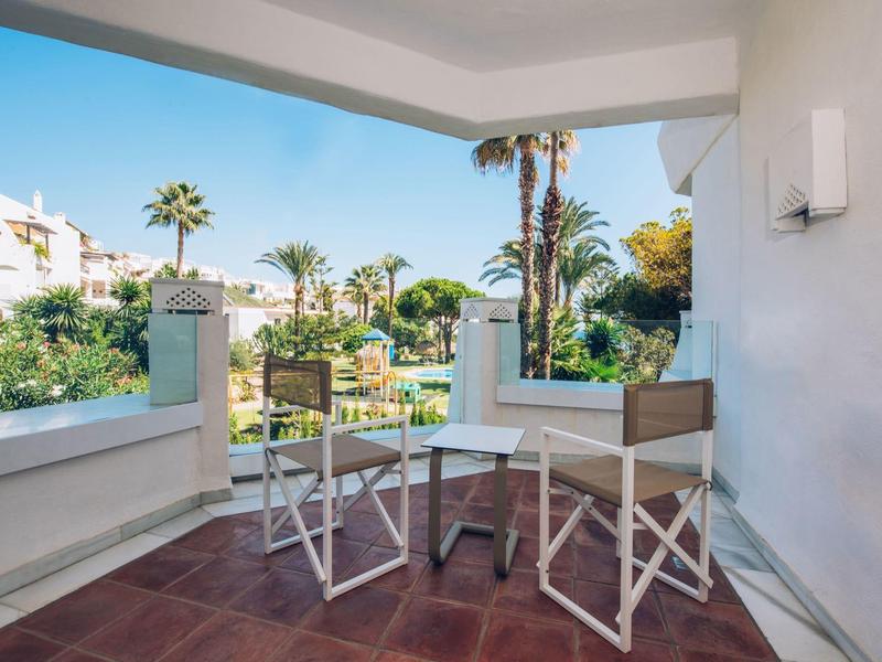 Balcony with two chairs and table, view of palm trees and white buildings under clear sky.