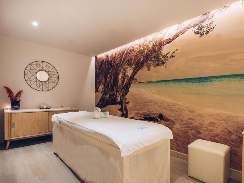 Wellness room with massage table, wooden sideboard, and tropical beach mural on the wall.