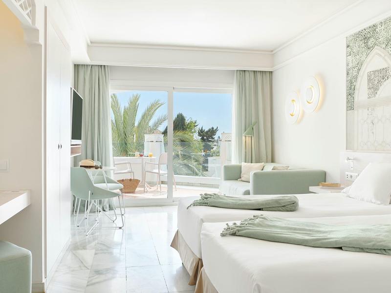Bright hotel room with two beds, balcony, and palm tree view.