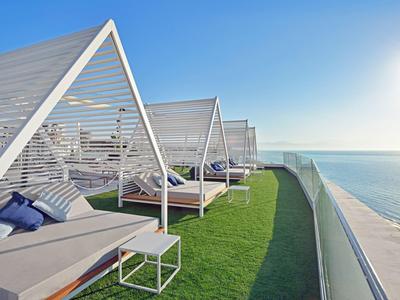 Modern lounge beds with cushions and shade on terrace overlooking ocean under clear sky.