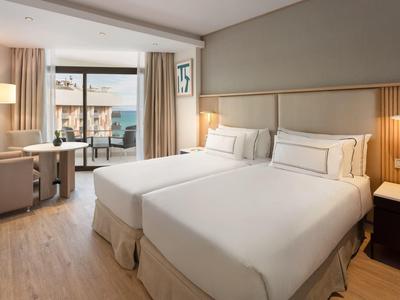 Bright hotel room with two single beds, desk, and balcony with sea view.