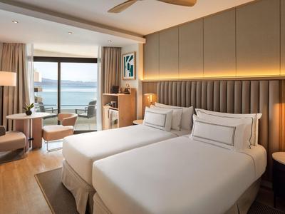 Modern hotel room with two beds, seating area, and ocean view through large windows.