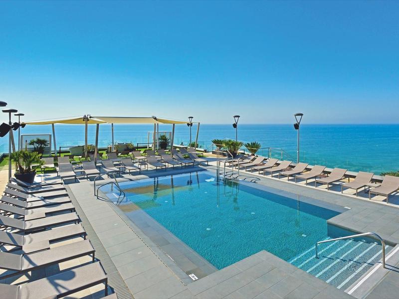 Modern pool with sun loungers and sea view under clear blue sky.