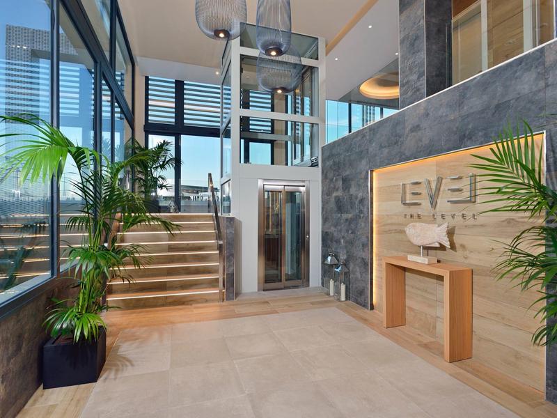 Modern hotel lobby with elevator, plants, and staircase with glass railing.