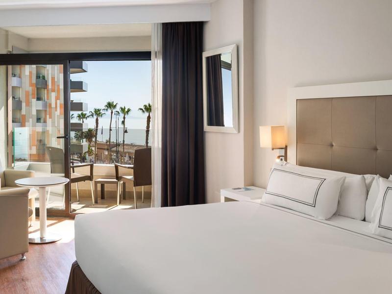 Bright hotel room with large bed, seating area, and view of balcony with palm trees.