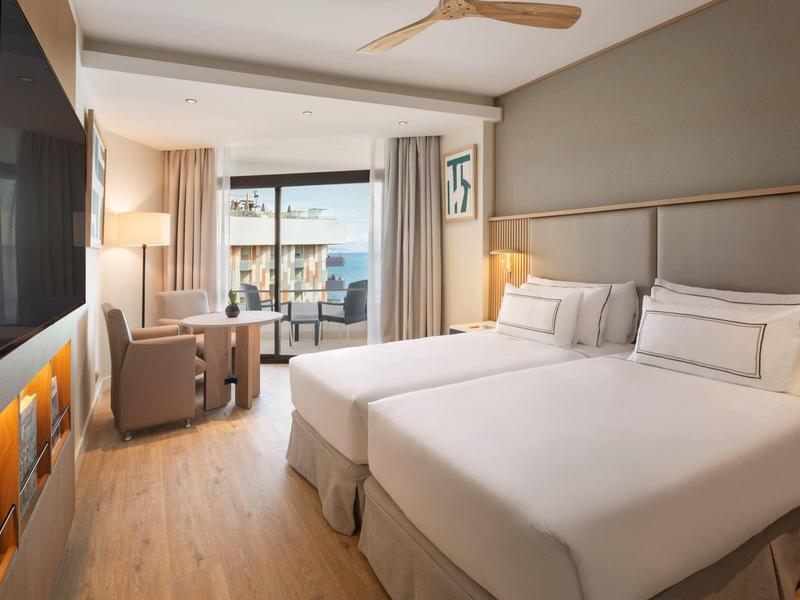 Bright hotel room with two beds, balcony, and modern decor featuring wooden flooring.