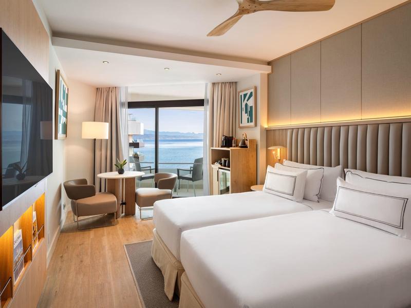 Modern hotel room with two beds, seating area, and sea view.