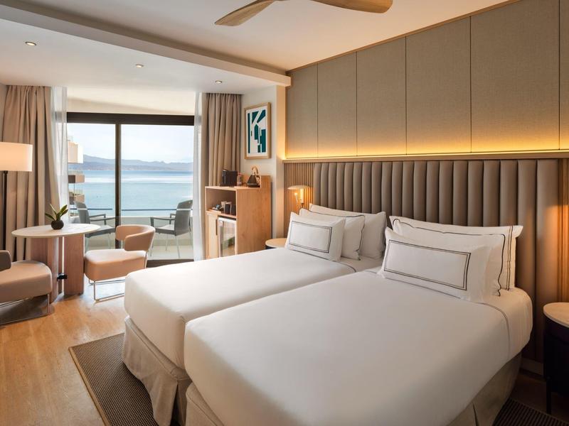 Modern hotel room with two beds, seating area, and ocean view through large windows.