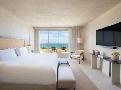 Helles Hotelzimmer mit großem Bett, TV, Stuhl und großem Fenster mit Meerblick.