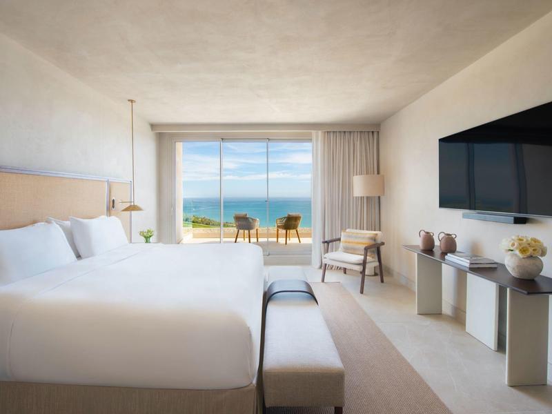 Helles Hotelzimmer mit großem Bett, TV, Stuhl und großem Fenster mit Meerblick.