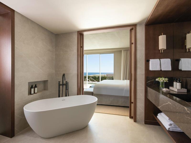 Moderne Badewanne und Waschtisch vor Schlafzimmer mit großer Fensterfront und Meerblick.