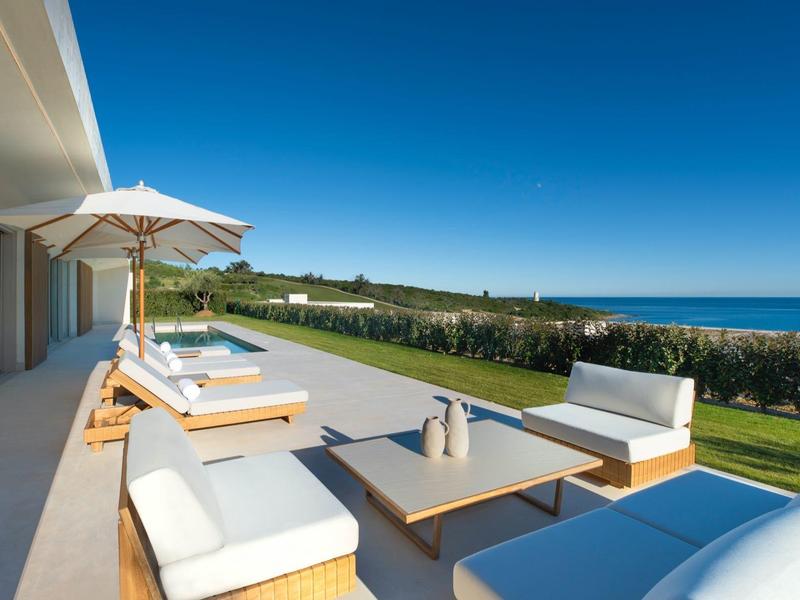 Moderne Terrasse mit weißen Loungemöbeln, Sonnenschirm, Blick auf grüne Wiesen und blauen Himmel.