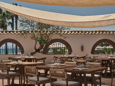 Area ristorante aperta con tavoli e sedie in legno, coperta da vele bianche, vista mare sullo sfondo.