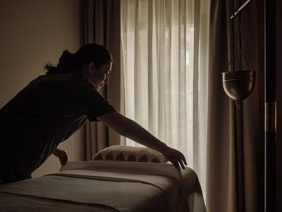 Persona che fa il letto in una stanza d'hotel poco illuminata con tende sullo sfondo.
