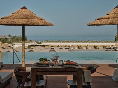 Un tavolo in legno apparecchiato con cibo e bevande accanto alla piscina sotto ombrelloni di paglia con vista mare.