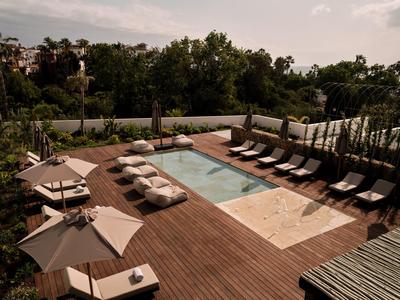 Piscina moderna con lettini e ombrelloni su terrazza in legno in contesto alberghiero verde.