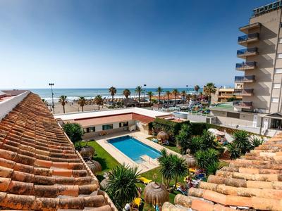Blick auf Swimmingpool und Promenade am Meer, umgeben von Gebäuden und Palmen bei klarem Himmel.