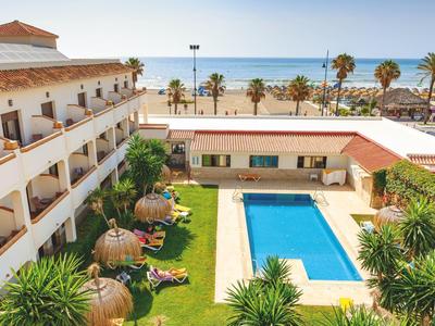 Hotel mit rechteckigem Pool, Liegestühlen und Palmen neben Strand und Meer im Hintergrund.