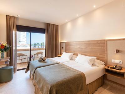 Helles Hotelzimmer mit Doppelbett, Holzkopfteil, Balkon und moderner Beleuchtung.