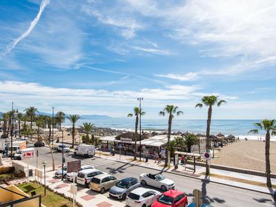 Strandpromenade mit parkenden Autos, Palmen und weitem Sandstrand unter blauem Himmel.