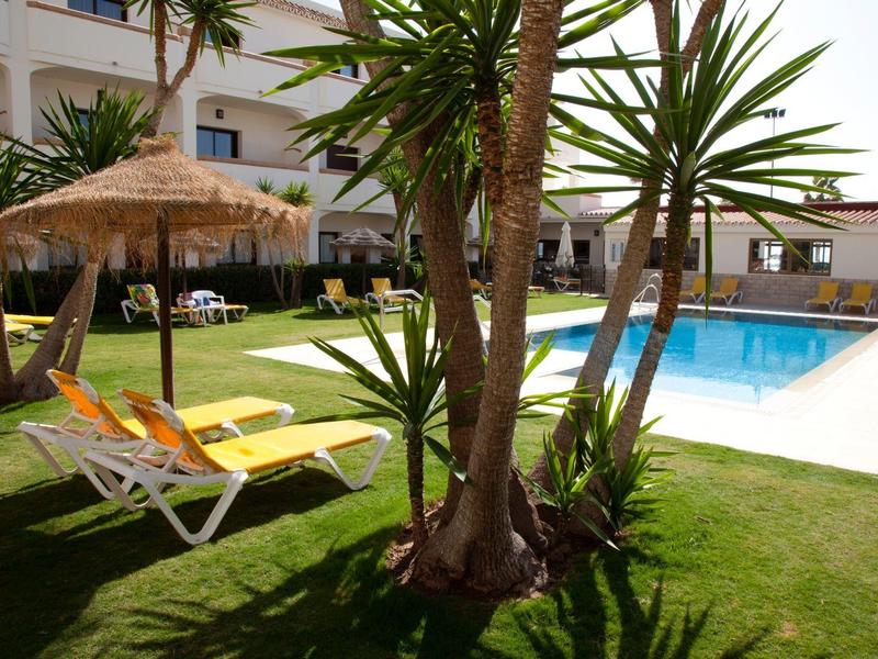 Hotel garden with pool, sun loungers, and palm trees on a sunny day.