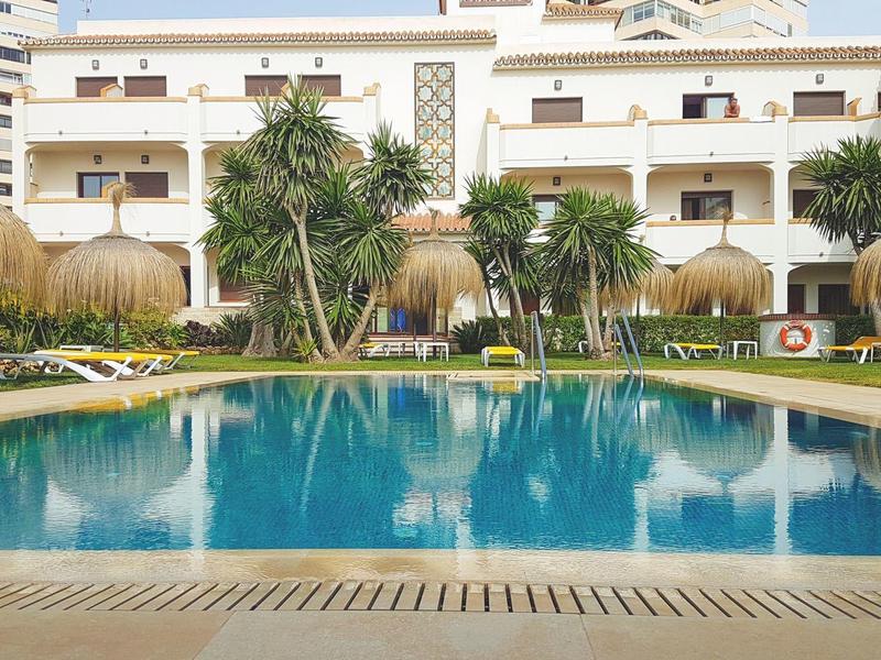 Large swimming pool with sun loungers and tropical plants in front of a hotel building.