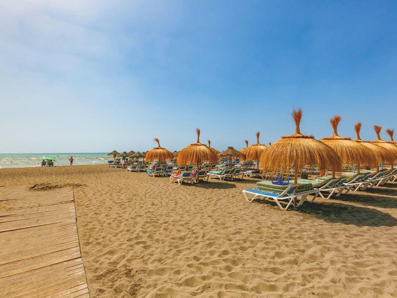 A quiet sandy beach with rows of empty loungers and closed umbrellas under a blue sky.