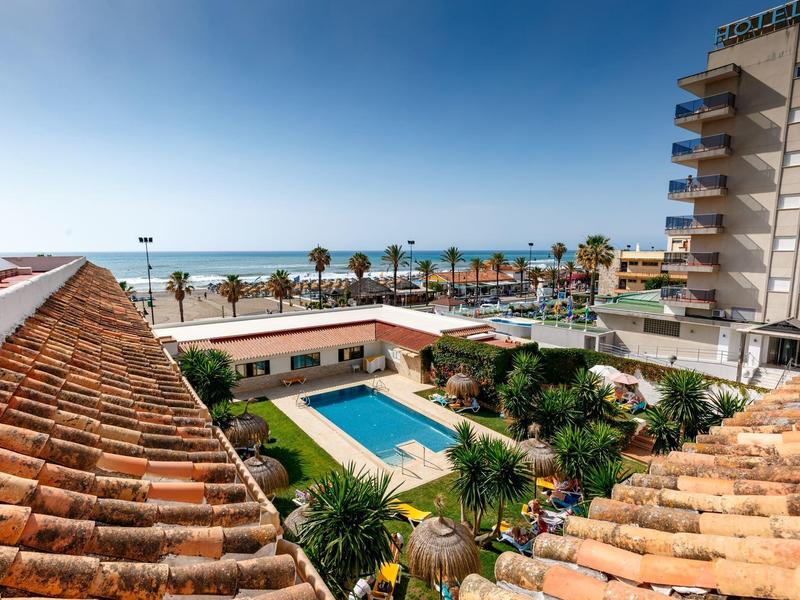 View of a hotel with pool and palm trees by the sea under a blue sky.