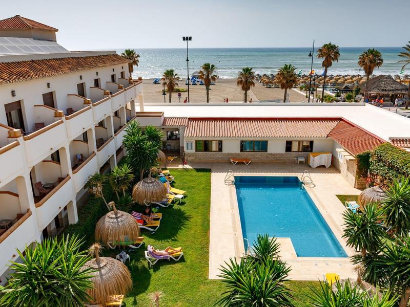 Hotel with pool and garden area, palm trees and sea view in the background.