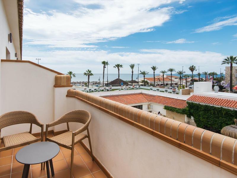 Balcony with chairs and table overlooking palm trees and buildings under cloudy sky.