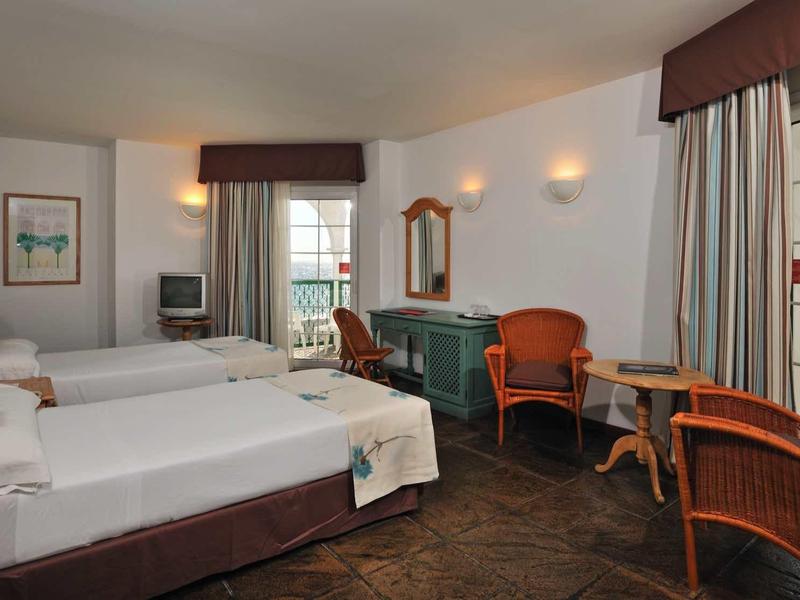 Hell eingerichtetes Hotelzimmer mit zwei Betten, Sessel, Tisch, Fernseher und großen Fenstern.