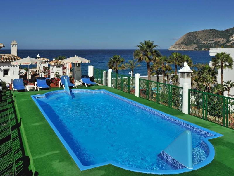 Schwimmbad mit blauem Wasser, grünem Rand, Palmen und Meer im Hintergrund unter klarem Himmel.