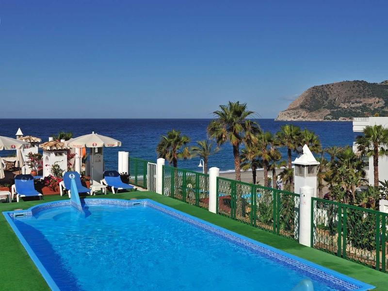 Schwimmbad mit blauer Umrandung, Palmen, Meer im Hintergrund und klarem Himmel.