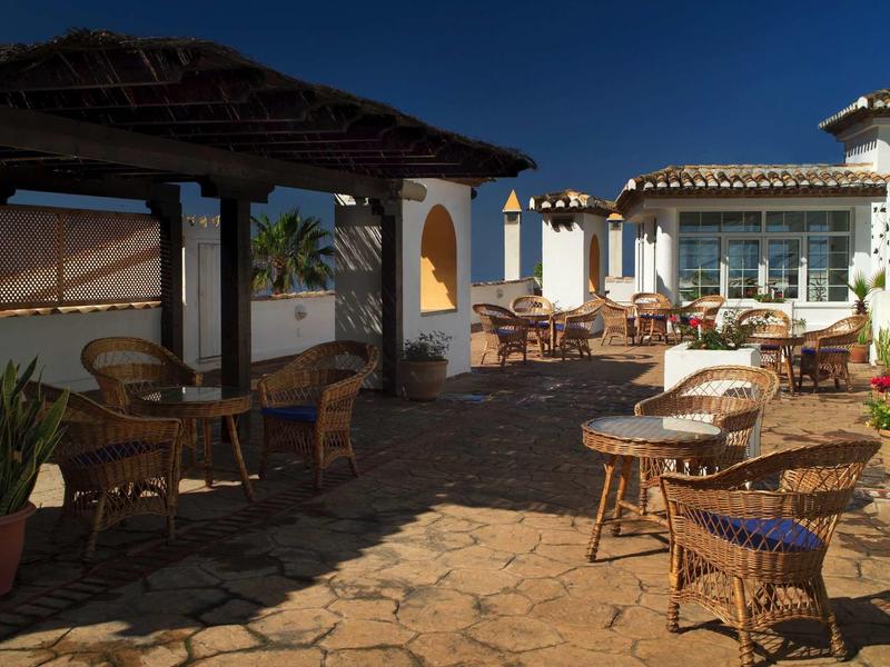 Terrasse mit Korbstühlen und Tischen unter blauem Himmel, Sonnenschutz und mediterranem Stil.