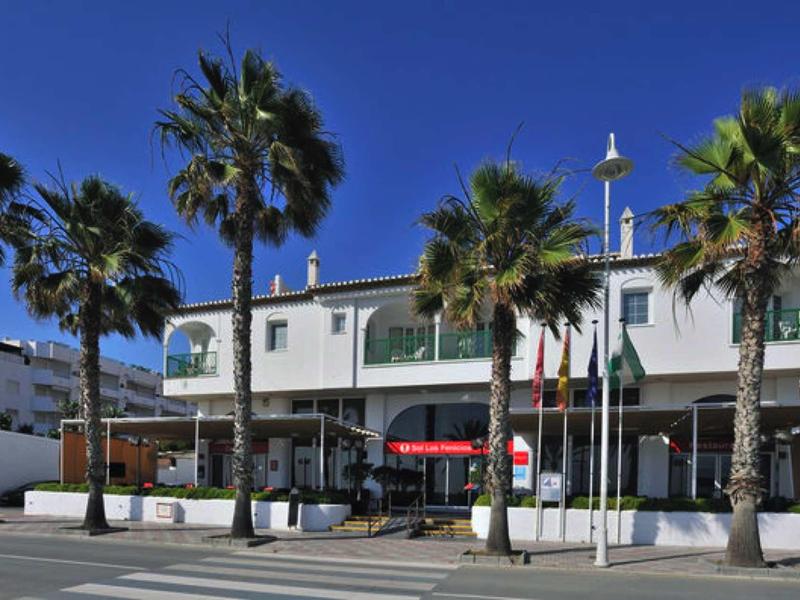 Gebäude mit vielen Palmen vor blauem Himmel und roten Sonnenschirmen auf Terrasse.