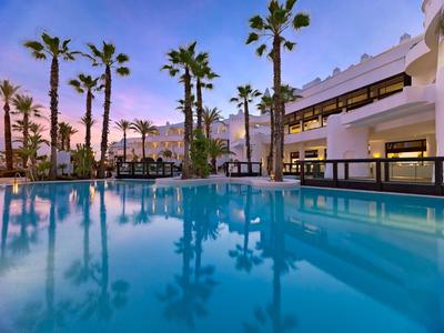 Großer blauer Pool bei Sonnenuntergang, umgeben von Palmen und weißem Hotelgebäude.