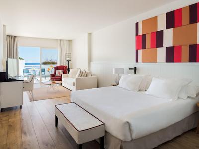 Helles Hotelzimmer mit großem Bett, Sofa, Stuhl, buntem Wandbild und Meerblick durch Fenster.