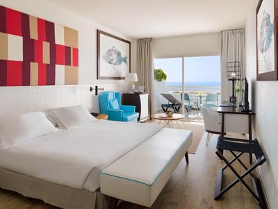 Helles Hotelzimmer mit Doppelbett, blauen Sesseln, Schreibtisch und Terrasse mit Meerblick.