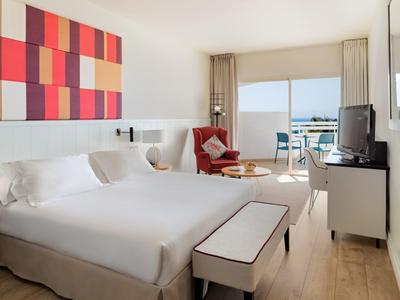 Modernes Schlafzimmer mit großem Bett, buntem Wandbild, rotem Sessel und Balkonblick auf Wasser.