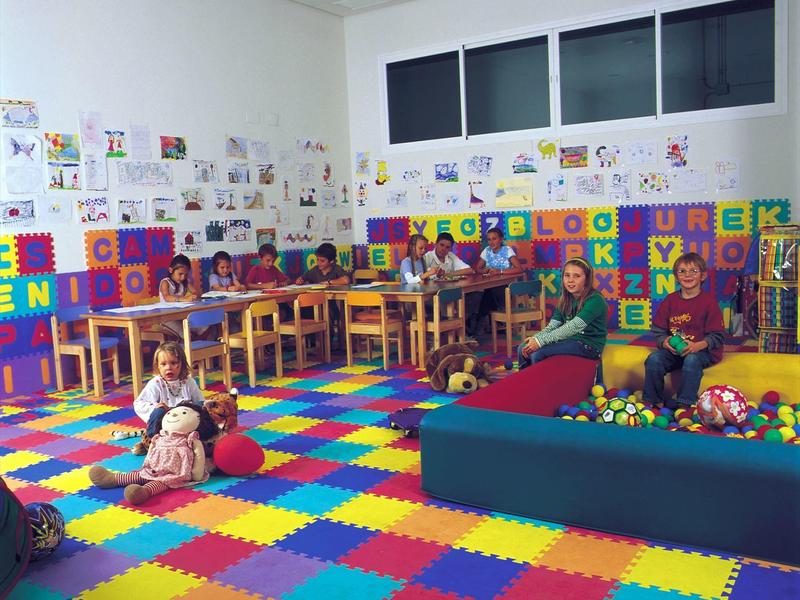 Bunter Kindergartenraum mit Spielteppich, Bällebad, Tischen, Stühlen und Kindern beim Spielen.