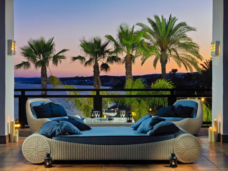 Gemütliche Lounge mit Polstermöbeln, Kissen und Kerzen, Palmen und Meer im Sonnenuntergang.