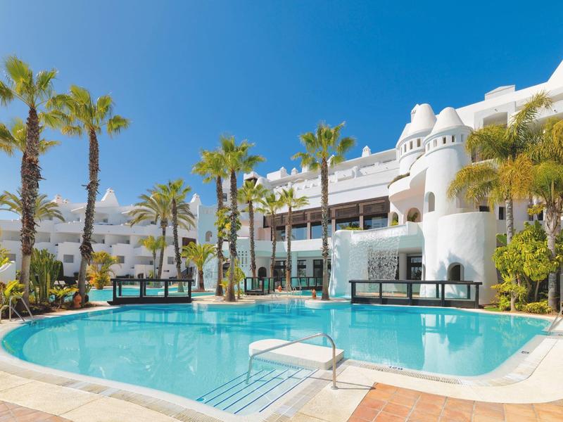 Großer Pool mit klar blauem Wasser, umgeben von weißen Gebäuden und Palmen unter blauem Himmel.