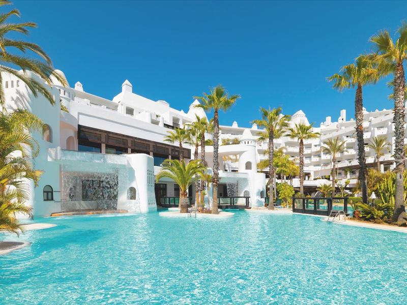 Großer blauer Pool vor weißem Hotel mit Palmen und blauem Himmel im Hintergrund.