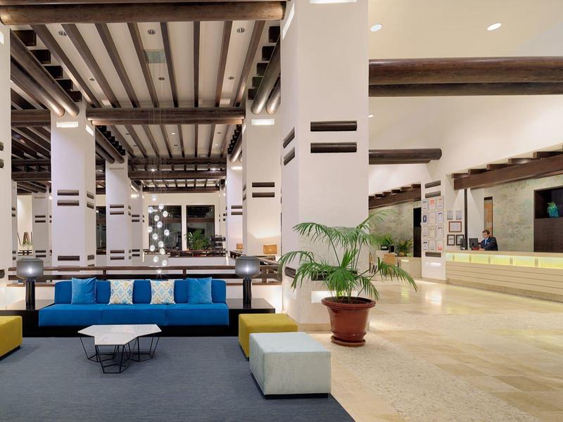 Große Hotellobby mit weißen Säulen, blauen Sofas, Holzdecken und einer Rezeption auf der rechten Seite.