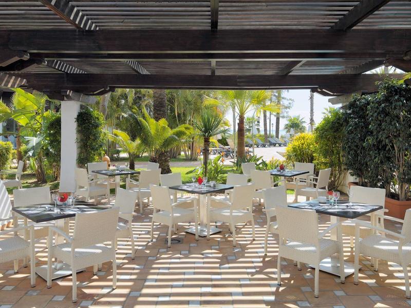 Außenbereich eines Cafés mit weißen Tischen und Stühlen unter Holzpergola, umgeben von Pflanzen.