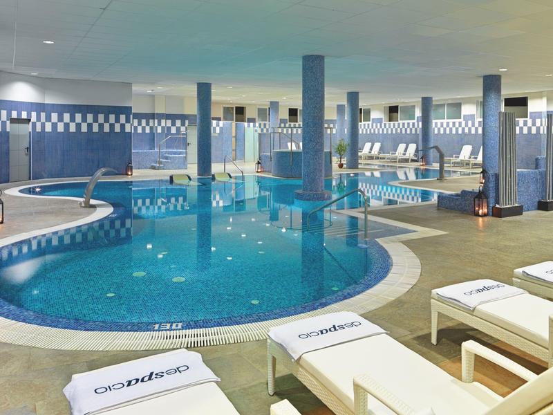 Innen-Schwimmbad mit blauem Wasser, Säulen und weißen Liegen am Beckenrand.