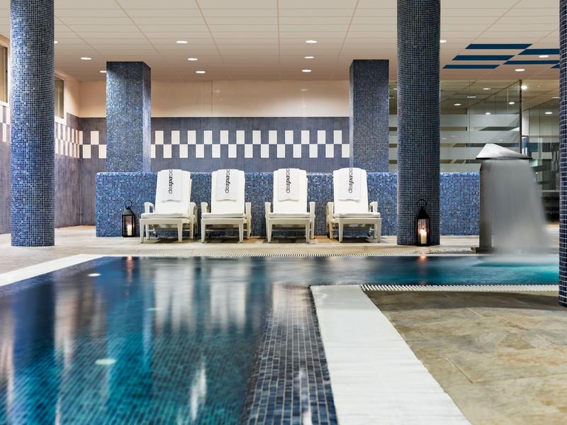 Innen-Schwimmbad mit blauem Becken, weißen Liegen und moderner Beleuchtung.