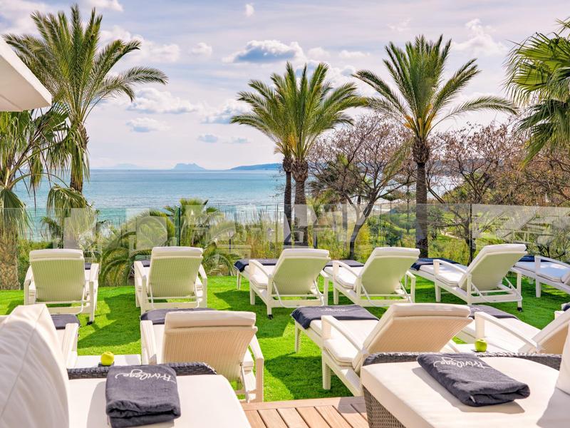 Grüne Liegewiese mit weißen Liegen und Handtüchern, Palmen und Meer im Hintergrund.