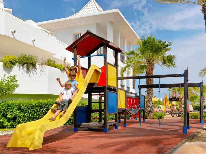 Kinder spielen auf gelber Rutsche und Klettergerüst neben weißem Gebäude und Palmen unter blauem Himmel.