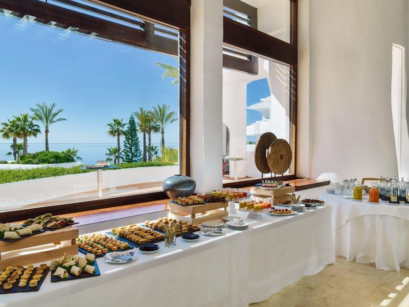 Buffet mit süßen und herzhaften Speisen vor großen Fenstern mit Meerblick und Palmen.